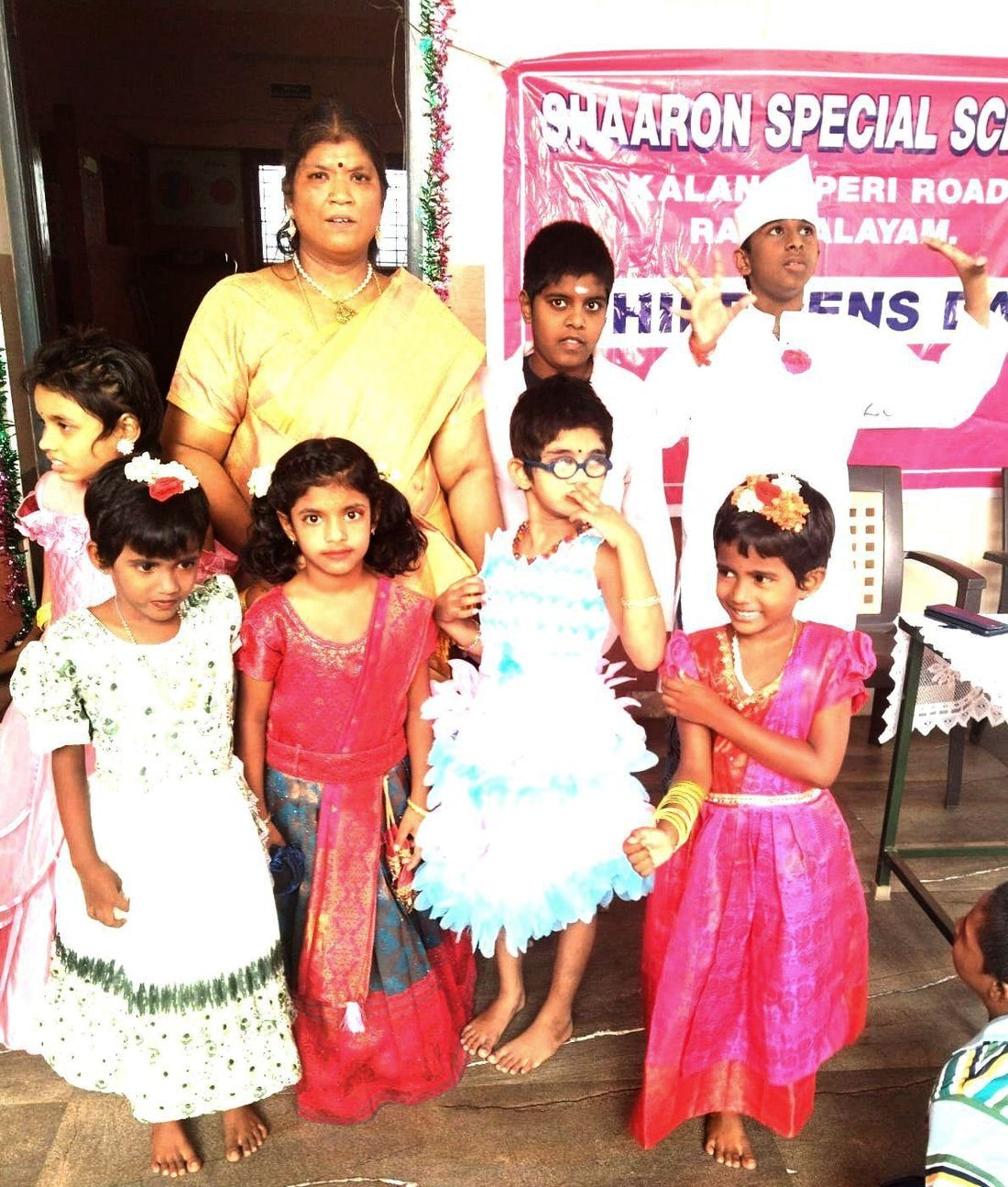 Young girls in party dresses stand with their teacher in front of the Children's Day banner