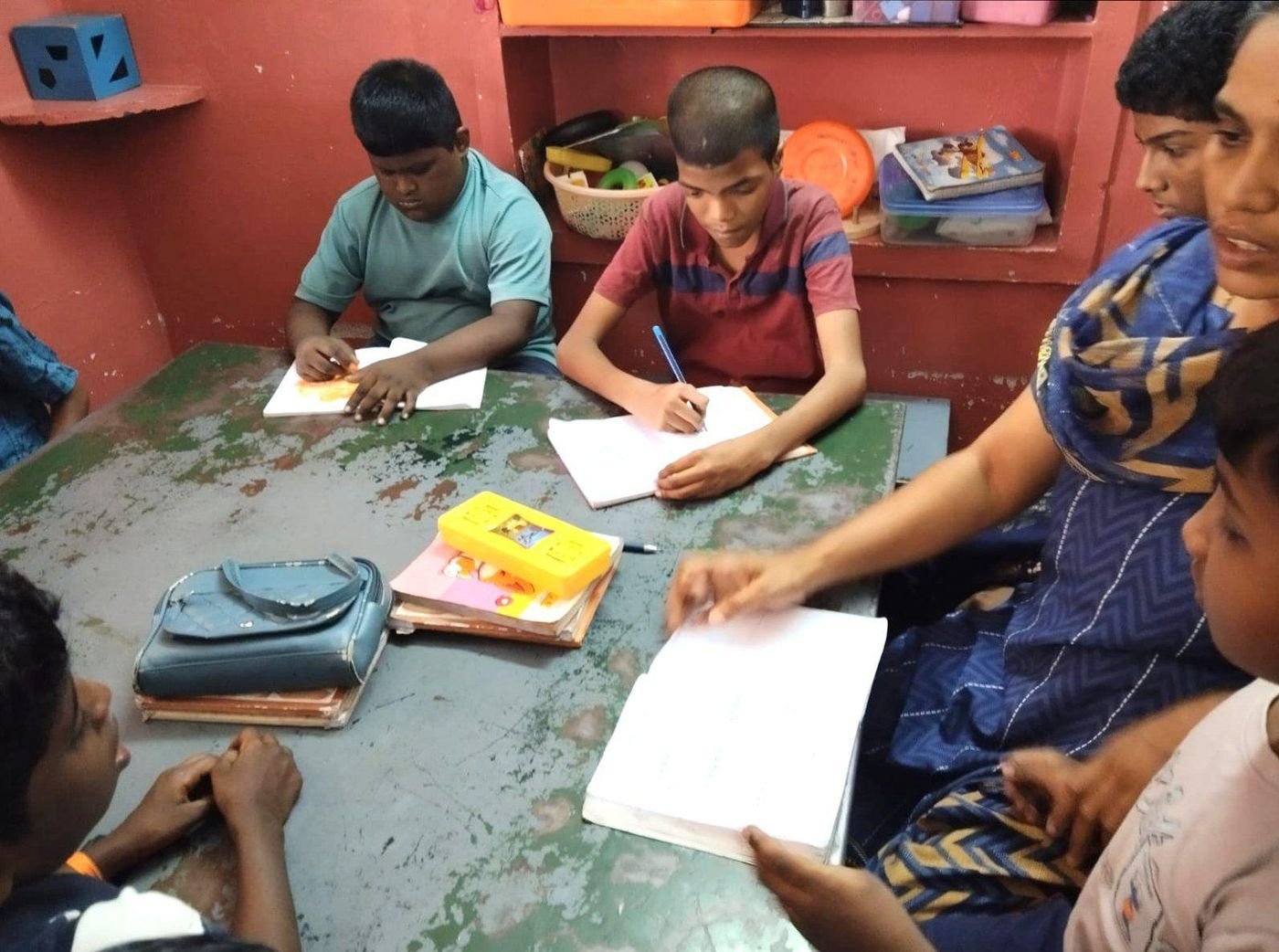 Students write in their notebooks at a classroom table while a teacher guides them