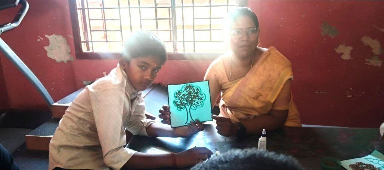 A student proudly shows the tree artwork he made, sitting beside his teacher