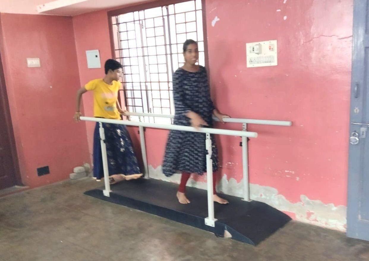 A student practises walking between parallel bars in the therapy room, with a staff member beside her
